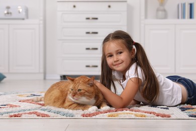 Photo of Smiling little girl petting cute ginger cat on carpet at home