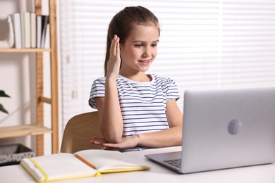 E-learning. Cute girl raising her hand to answer during online lesson at table indoors