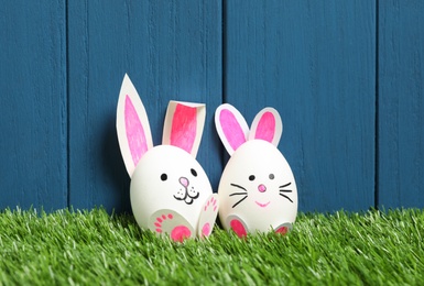 Photo of Bright Easter eggs as cute bunnies on green grass against blue wooden background
