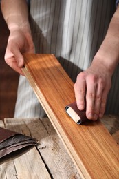 Man polishing wooden plank with sandpaper at table indoors, closeup