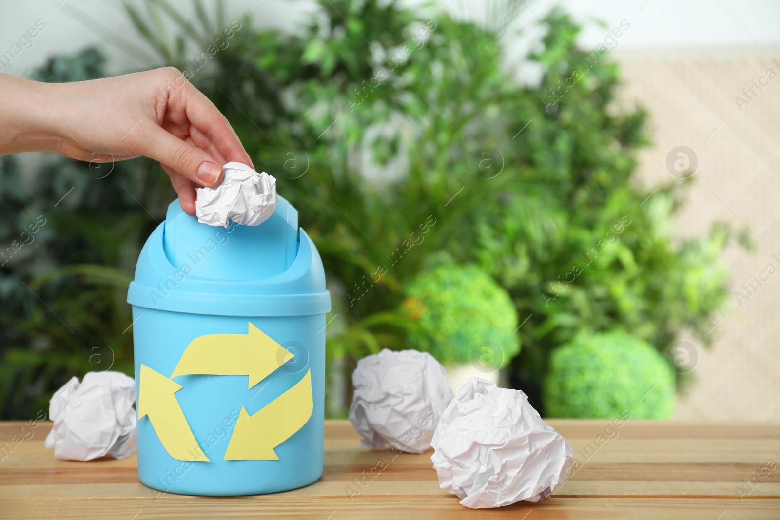 Photo of Woman throwing crumpled paper into trash bin with recycling symbol at wooden table, space for text