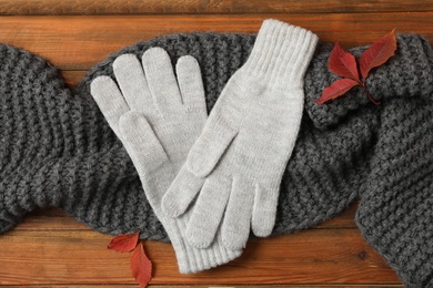 Stylish woolen gloves, scarf and dry leaves on wooden table, flat lay