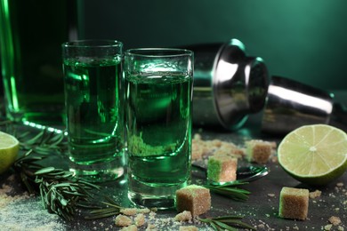 Absinthe in shot glasses, spoon, brown sugar, lime and rosemary on gray table, closeup. Alcoholic drink