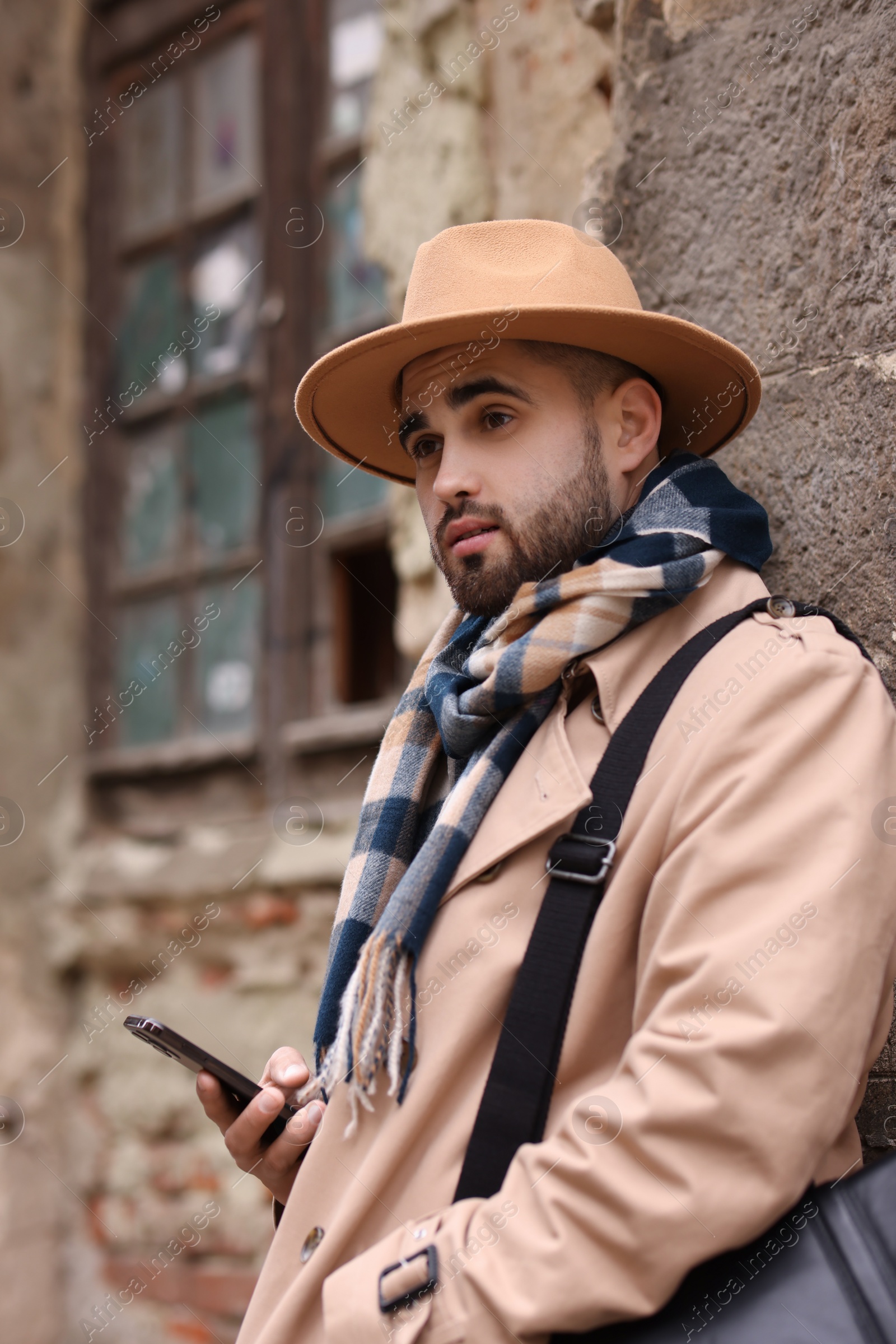 Photo of Handsome man in warm scarf with smartphone near wall outdoors