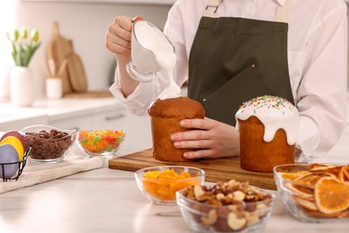 Photo of Woman decorating traditional Easter cake with glaze at white marble table in kitchen, closeup