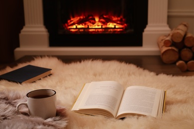 Cup of hot tea and books on faux fur near fireplace at home. Cozy atmosphere