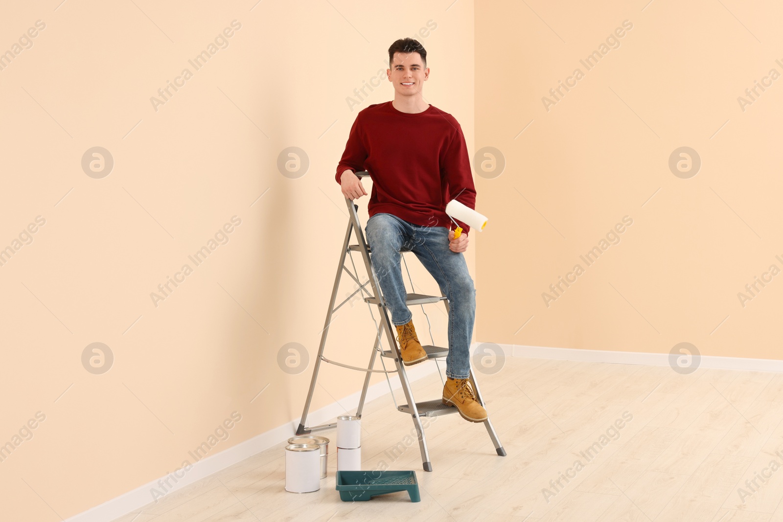 Photo of Young handsome man with roller on metal stepladder indoors. Room renovation