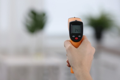 Closeup view of woman with infrared thermometer on blurred background, space for text. Checking temperature during Covid-19 pandemic