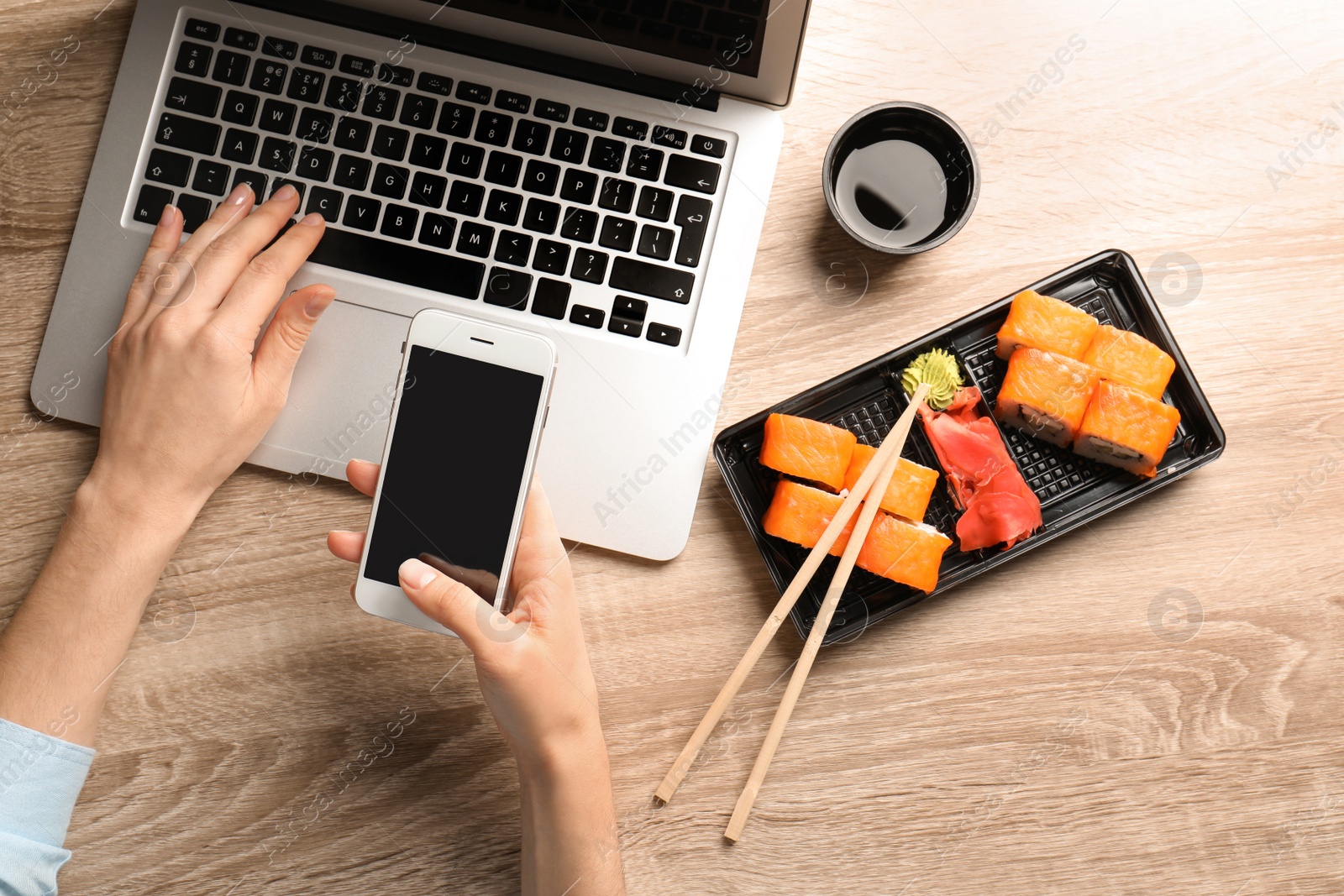 Photo of Woman with smartphone, laptop and tasty sushi rolls at wooden table, space for text. Food delivery