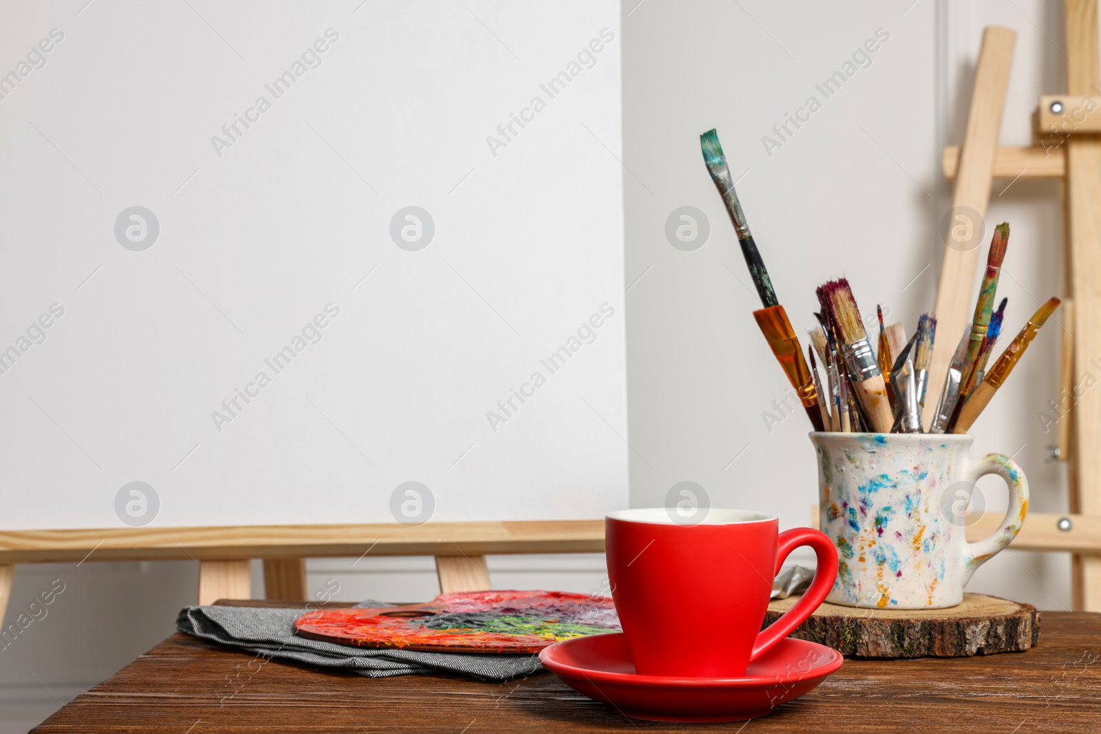 Photo of Easel with blank canvas, cup of drink and different art supplies on wooden table near white wall