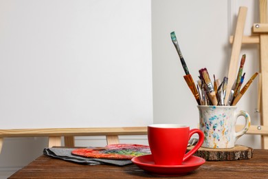 Easel with blank canvas, cup of drink and different art supplies on wooden table near white wall