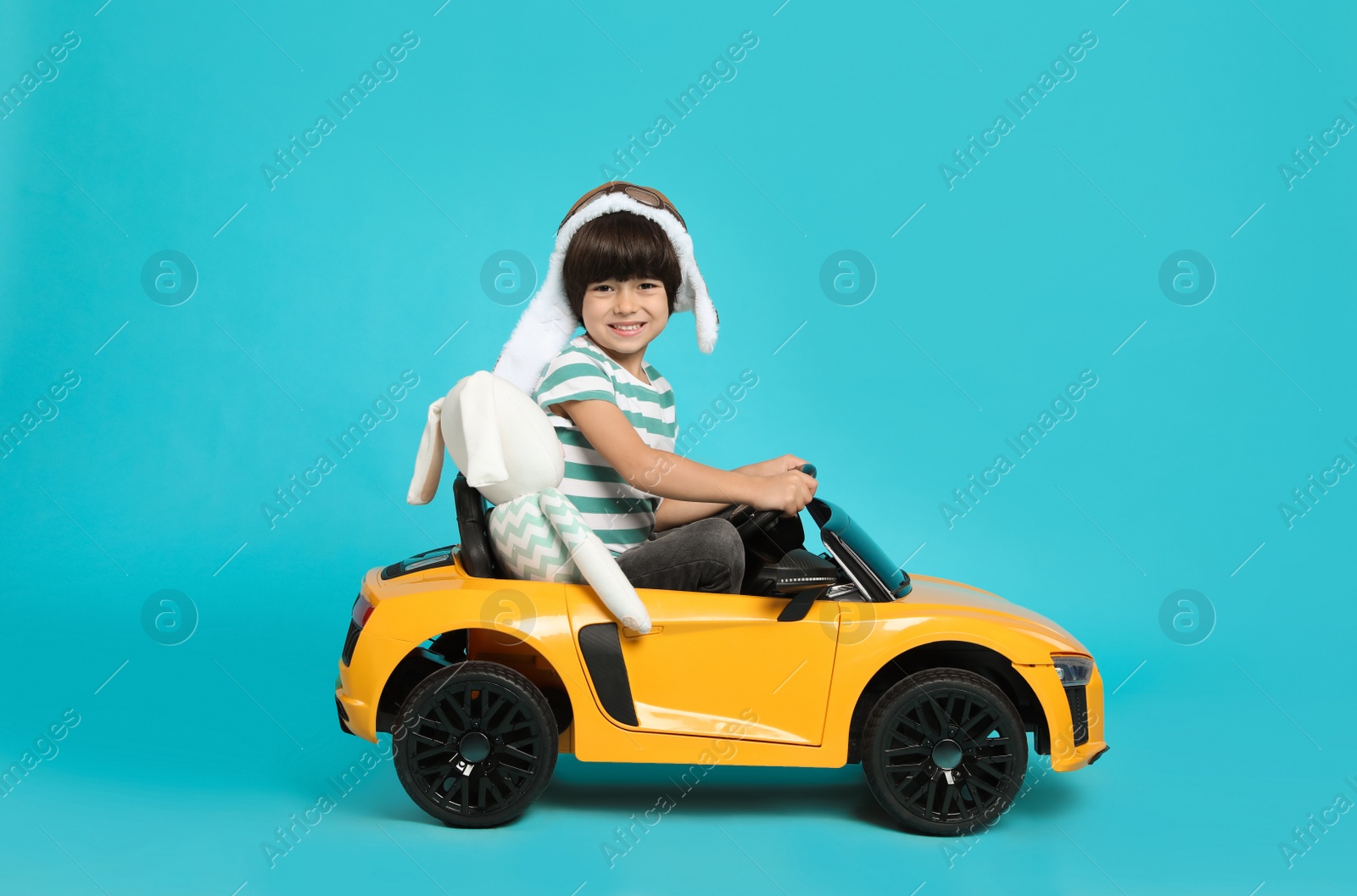 Photo of Cute little boy with toy bunny driving children's car on light blue background