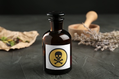 Photo of Glass bottle of poison with warning sign and herb on grey stone table