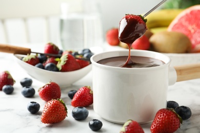 Photo of Dipping strawberry into fondue pot with chocolate on white marble table