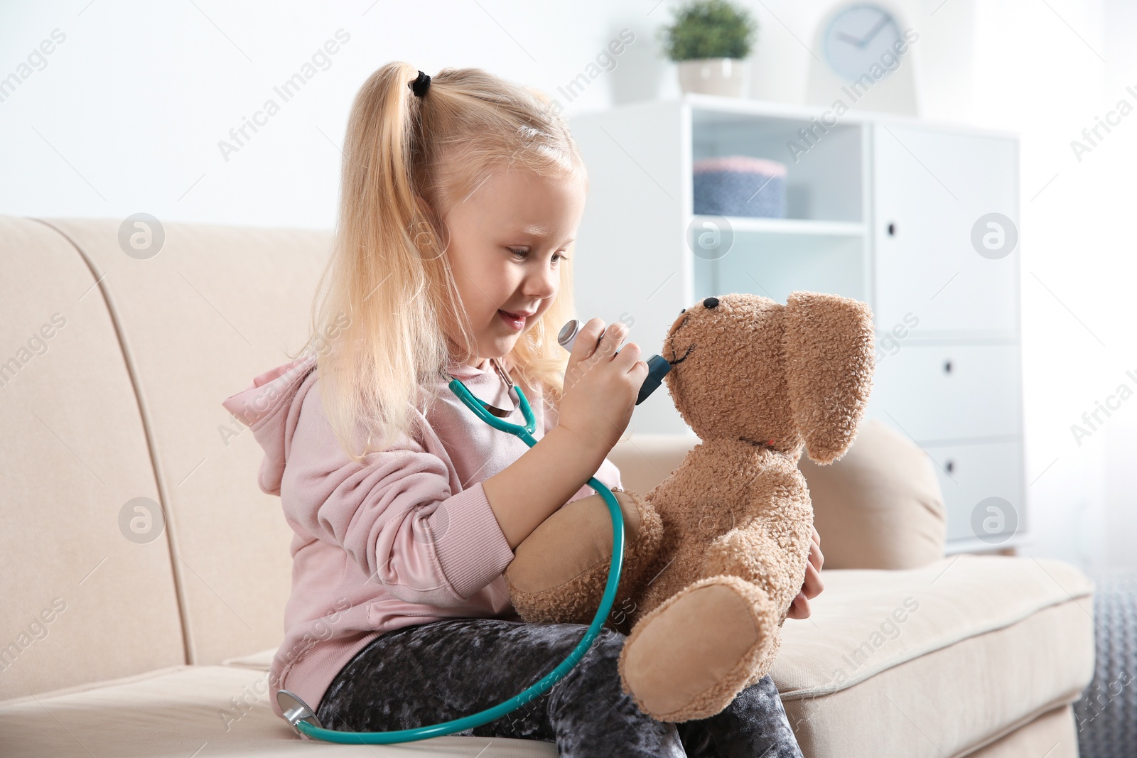 Photo of Cute child imagining herself as doctor while playing with toy bunny on couch at home