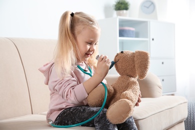 Photo of Cute child imagining herself as doctor while playing with toy bunny on couch at home