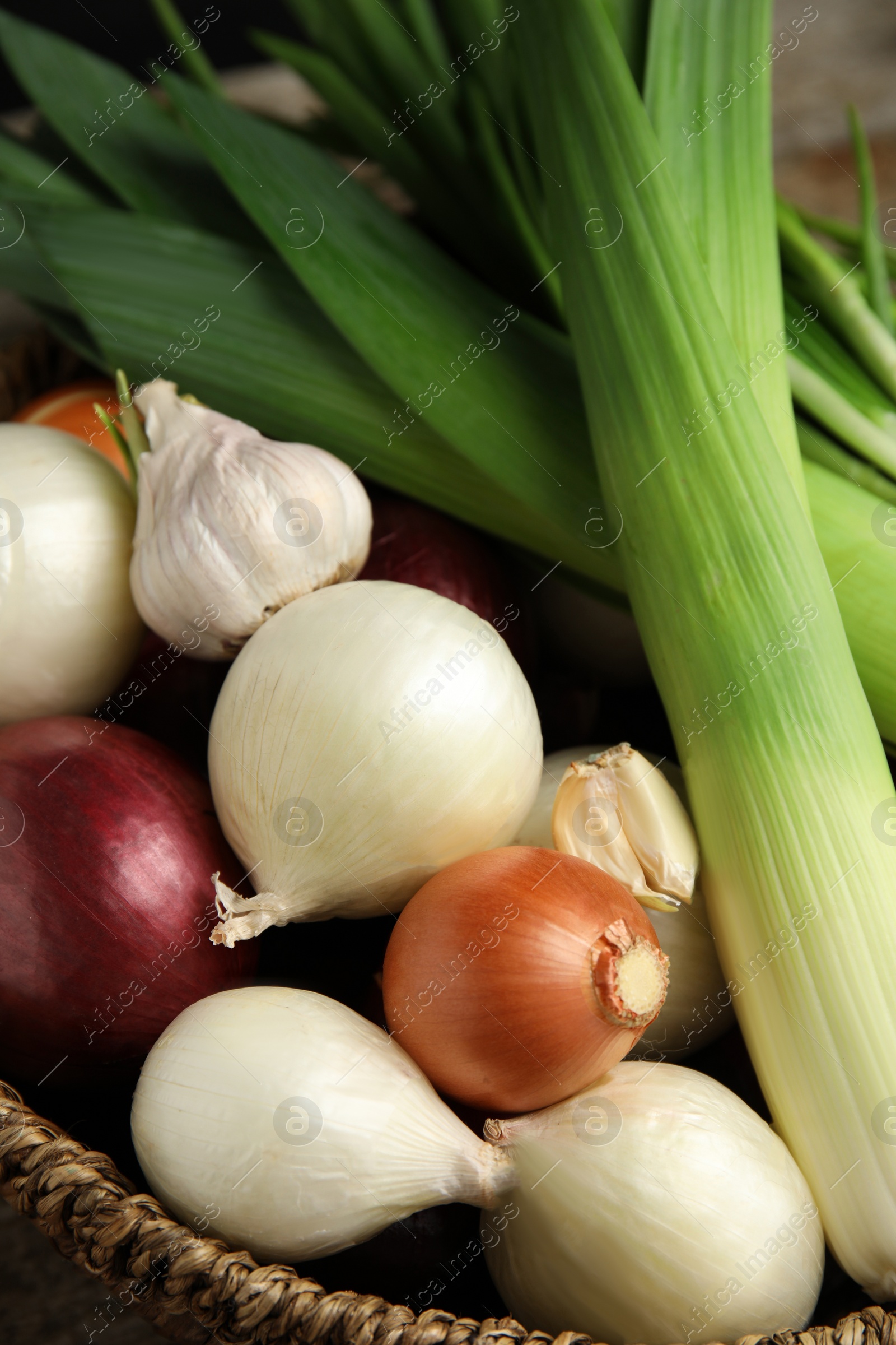 Photo of Fresh onion bulbs, leeks and garlic in wicker basket, closeup