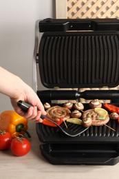 Photo of Woman cooking homemade sausages with bell peppers and mushrooms on electric grill at wooden table, closeup