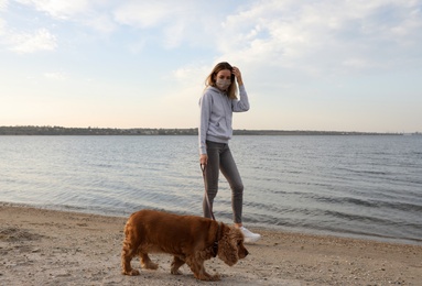 Woman in protective mask with English Cocker Spaniel on beach. Walking dog during COVID-19 pandemic