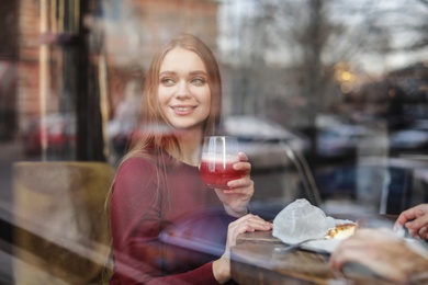 Photo of Pretty young woman with cocktail and cake at table in cafe, view from outdoors through window