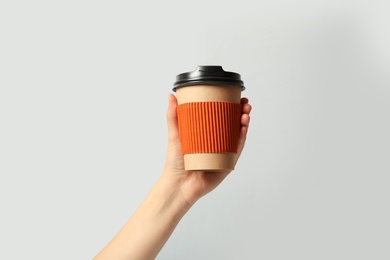 Woman holding takeaway paper coffee cup with cardboard sleeve on light grey background, closeup