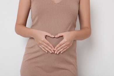 Pregnant woman in beige dress making heart with hands on white background, closeup