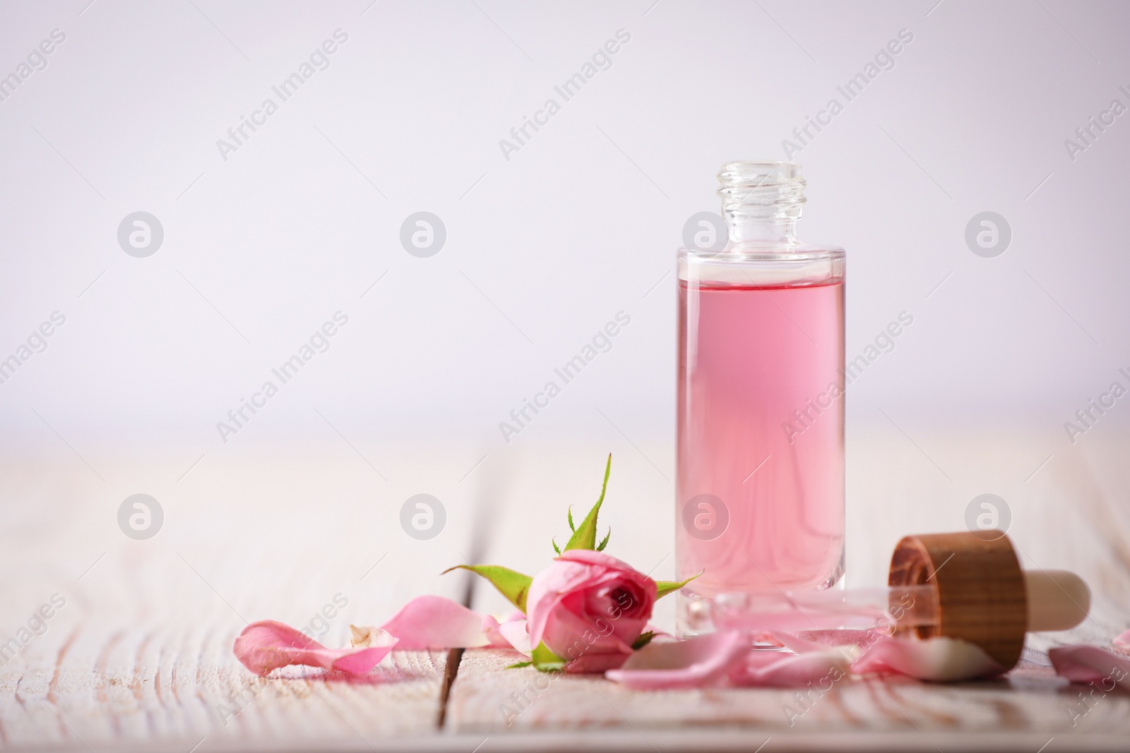 Photo of Bottle of essential rose oil and flowers on white wooden table against light background. Space for text