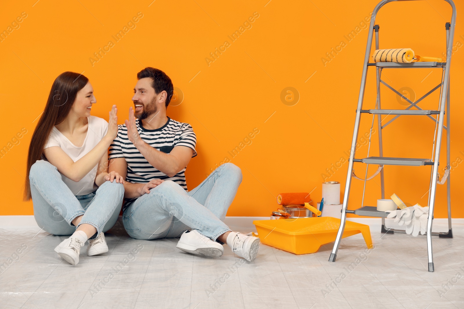 Photo of Happy designers giving high five on floor near freshly painted orange wall indoors