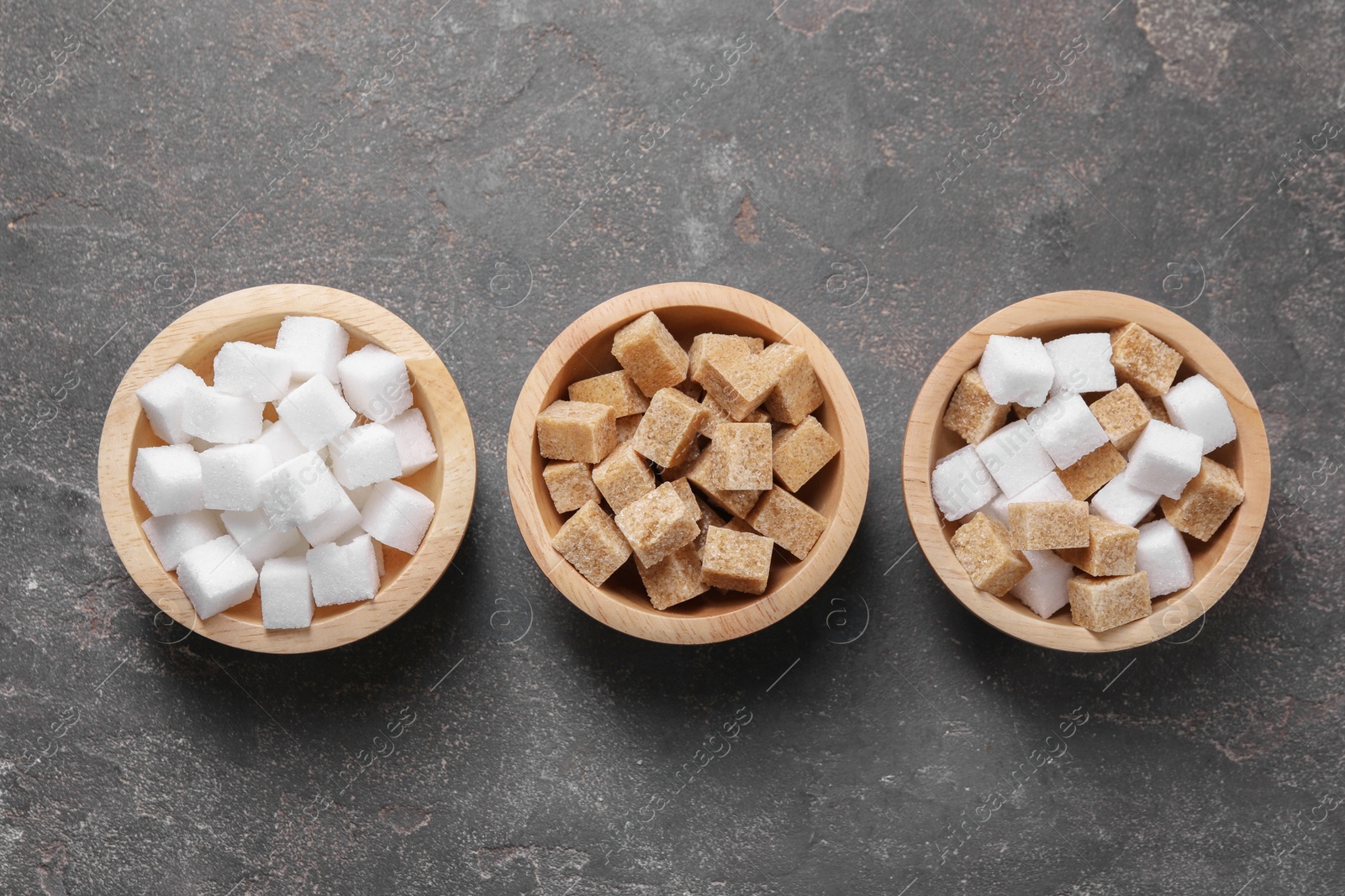 Photo of Different sugar cubes in bowls on gray textured table, flat lay