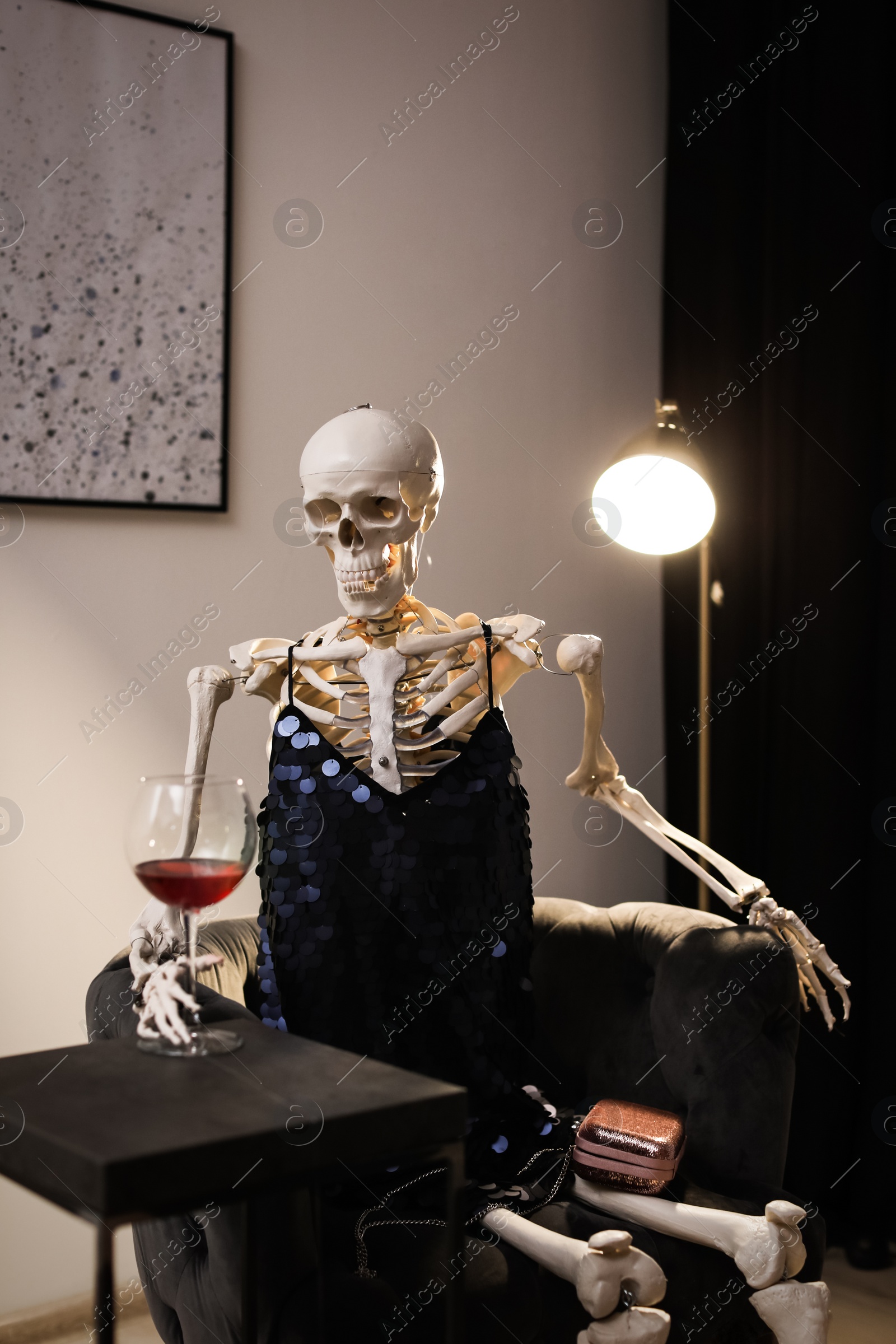 Photo of Skeleton in dress with wine sitting at table indoors