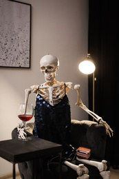 Photo of Skeleton in dress with wine sitting at table indoors