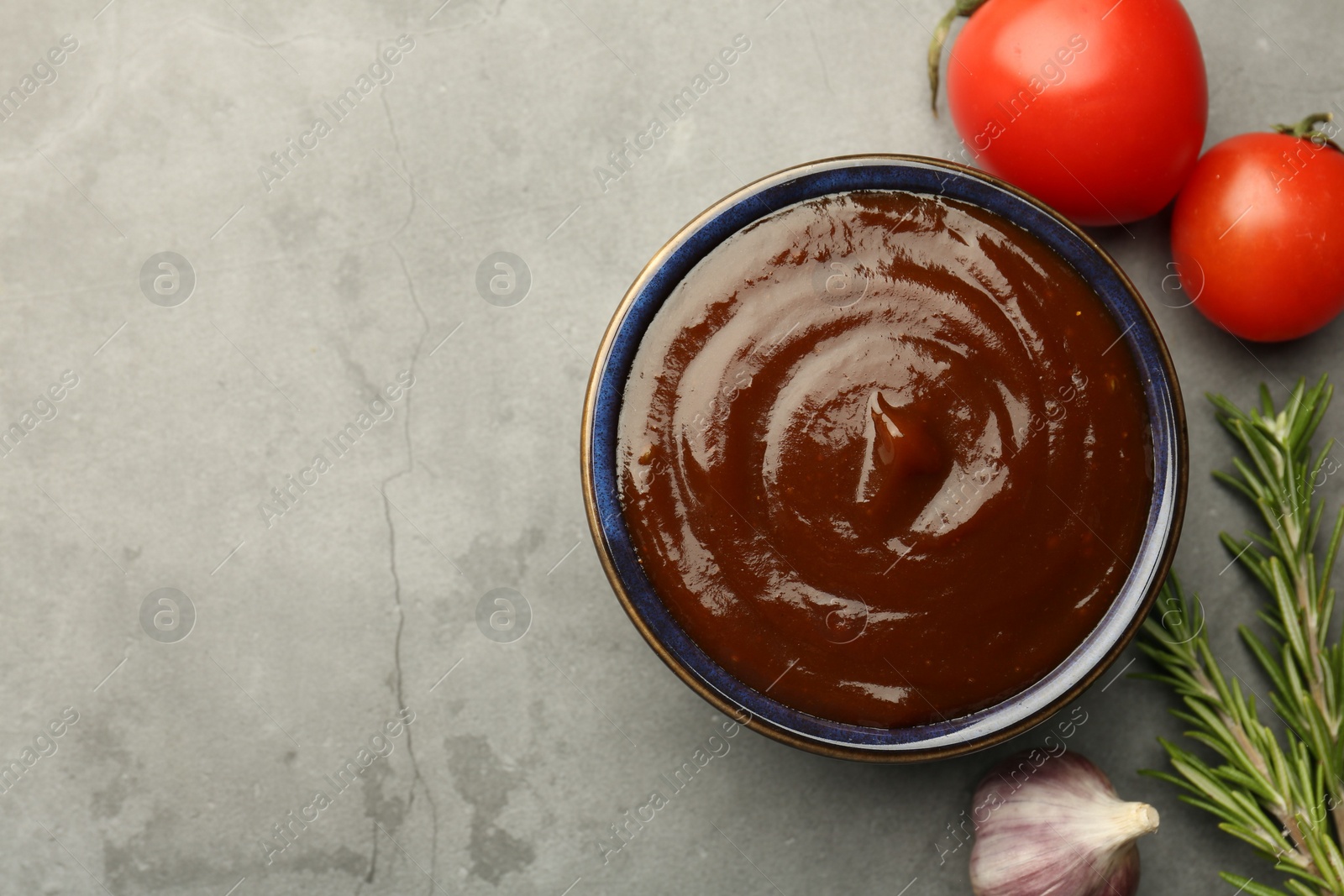 Photo of Tasty barbeque sauce in bowl, rosemary, garlic and tomato on grey textured table, flat lay. Space for text