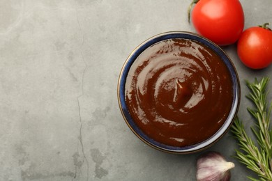 Photo of Tasty barbeque sauce in bowl, rosemary, garlic and tomato on grey textured table, flat lay. Space for text
