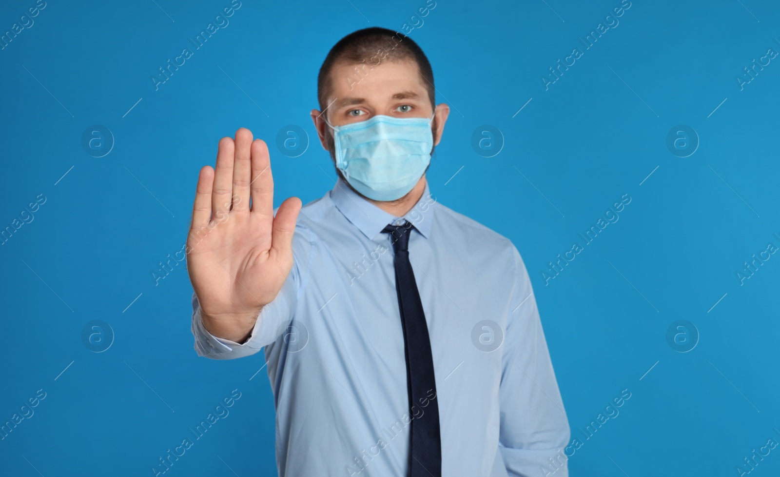 Photo of Man in protective mask showing stop gesture on light blue background. Prevent spreading of coronavirus