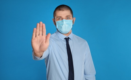 Photo of Man in protective mask showing stop gesture on light blue background. Prevent spreading of coronavirus