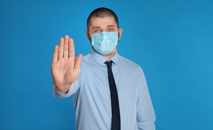 Man in protective mask showing stop gesture on light blue background. Prevent spreading of coronavirus