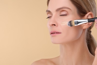 Woman applying foundation on face with brush against beige background. Space for text