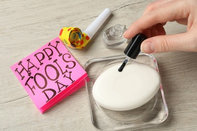 Photo of Woman applying transparent nail polish onto soap bar near paper with words Happy Fool's Day at wooden table, closeup