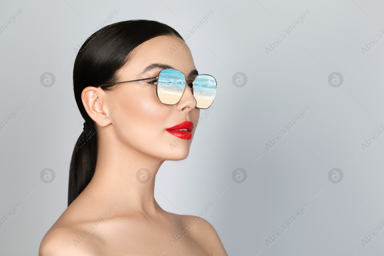 Image of Attractive woman in stylish sunglasses on light grey background. Sandy beach and sea reflecting in lenses. Space for text