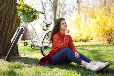 Photo of Beautiful woman sitting on green grass near bicycle with bouquet of yellow tulips outdoors. Spring day