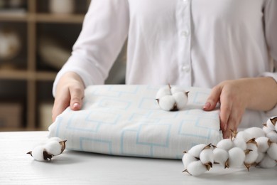 Photo of Woman taking bed sheet and cotton flowers on white wooden table in room, closeup