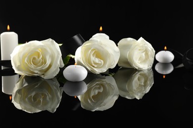 Photo of White roses and burning candles on black mirror surface in darkness. Funeral symbols