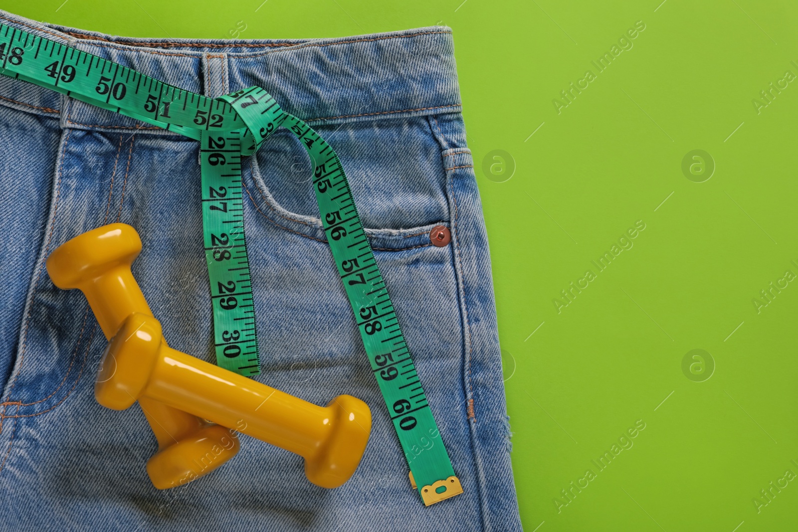 Photo of Jeans, dumbbells and measuring tape on light green background, flat lay with space for text. Weight loss concept