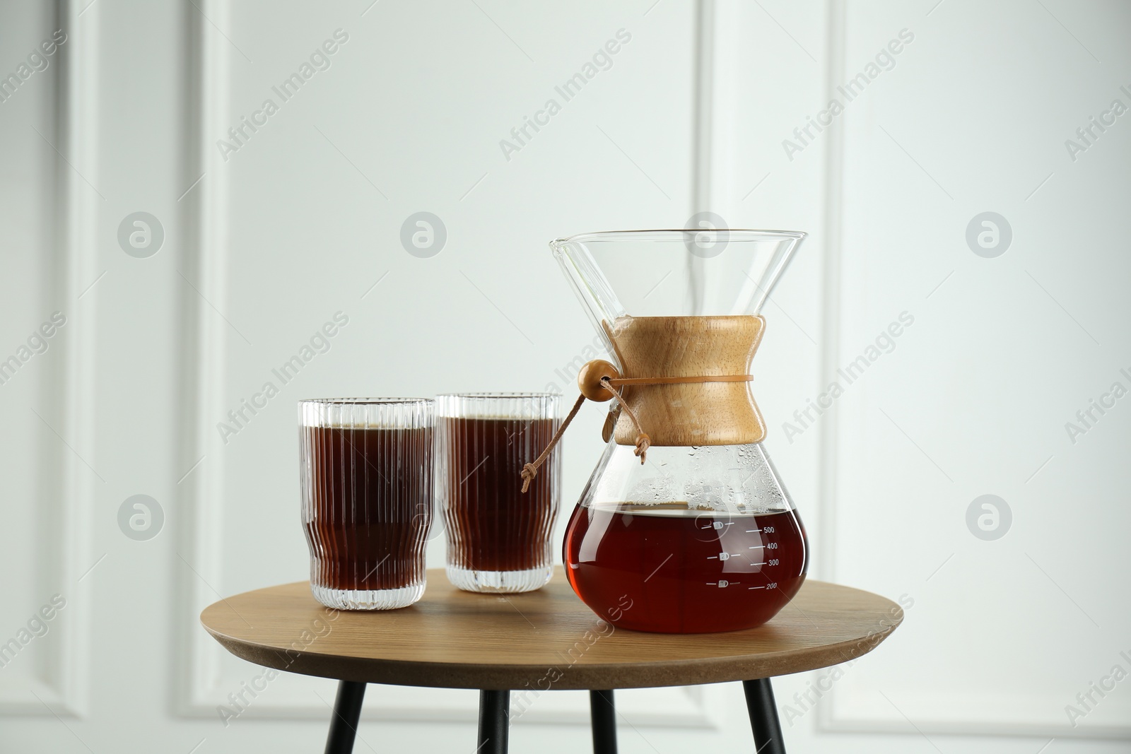 Photo of Glass chemex coffeemaker and glasses of coffee on wooden table against white wall