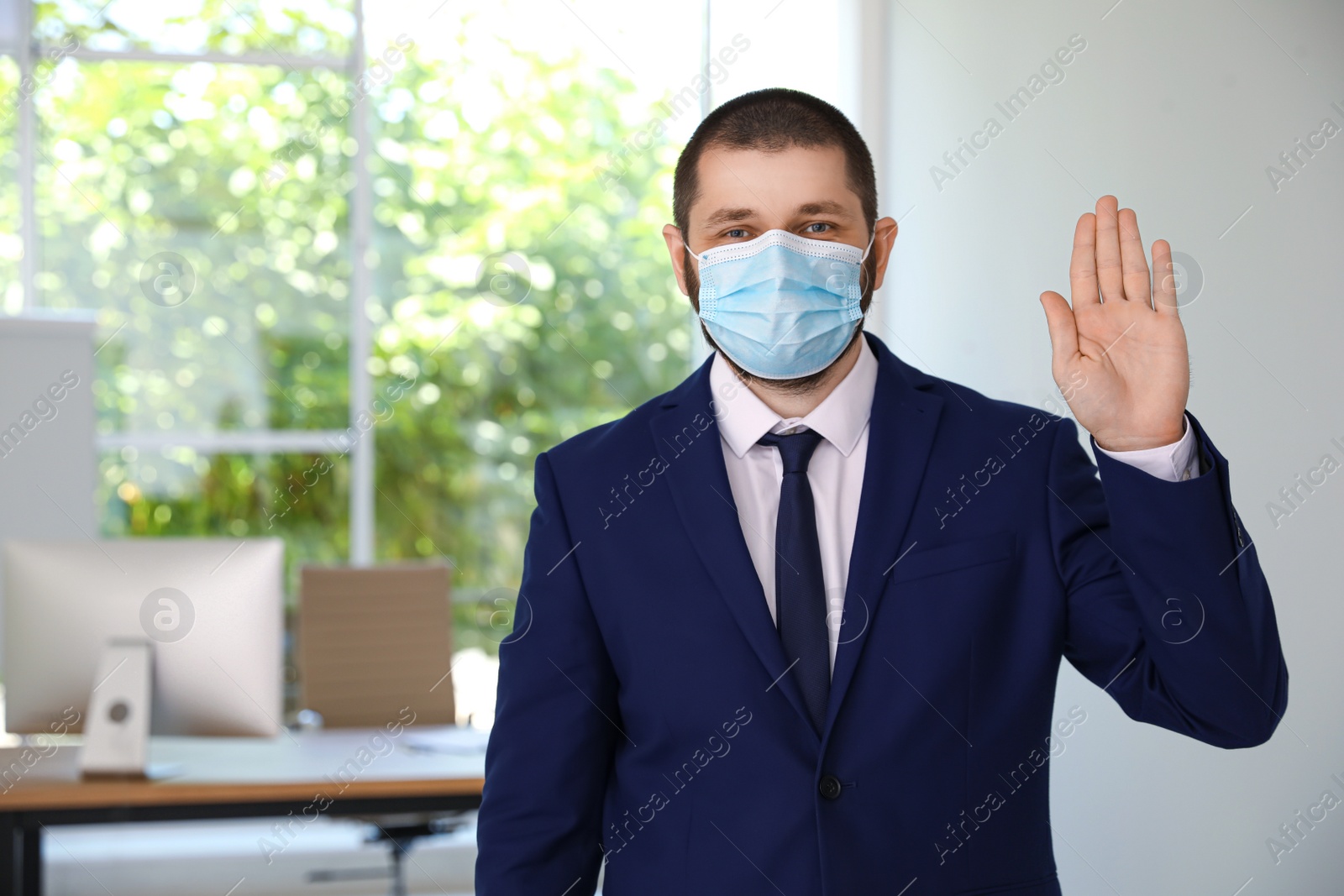 Photo of Man in protective face mask showing hello gesture in office. Keeping social distance during coronavirus pandemic