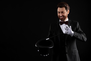 Happy magician showing magic trick with top hat on black background, space for text