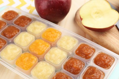 Photo of Different purees in ice cube tray and fresh apple fruits on wooden table, closeup. Ready for freezing