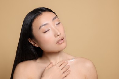 Photo of Beautiful young Asian woman applying body cream on shoulder against beige background, space for text