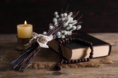 Rosary beads, Bible, burning candle and willow branches on wooden table, closeup