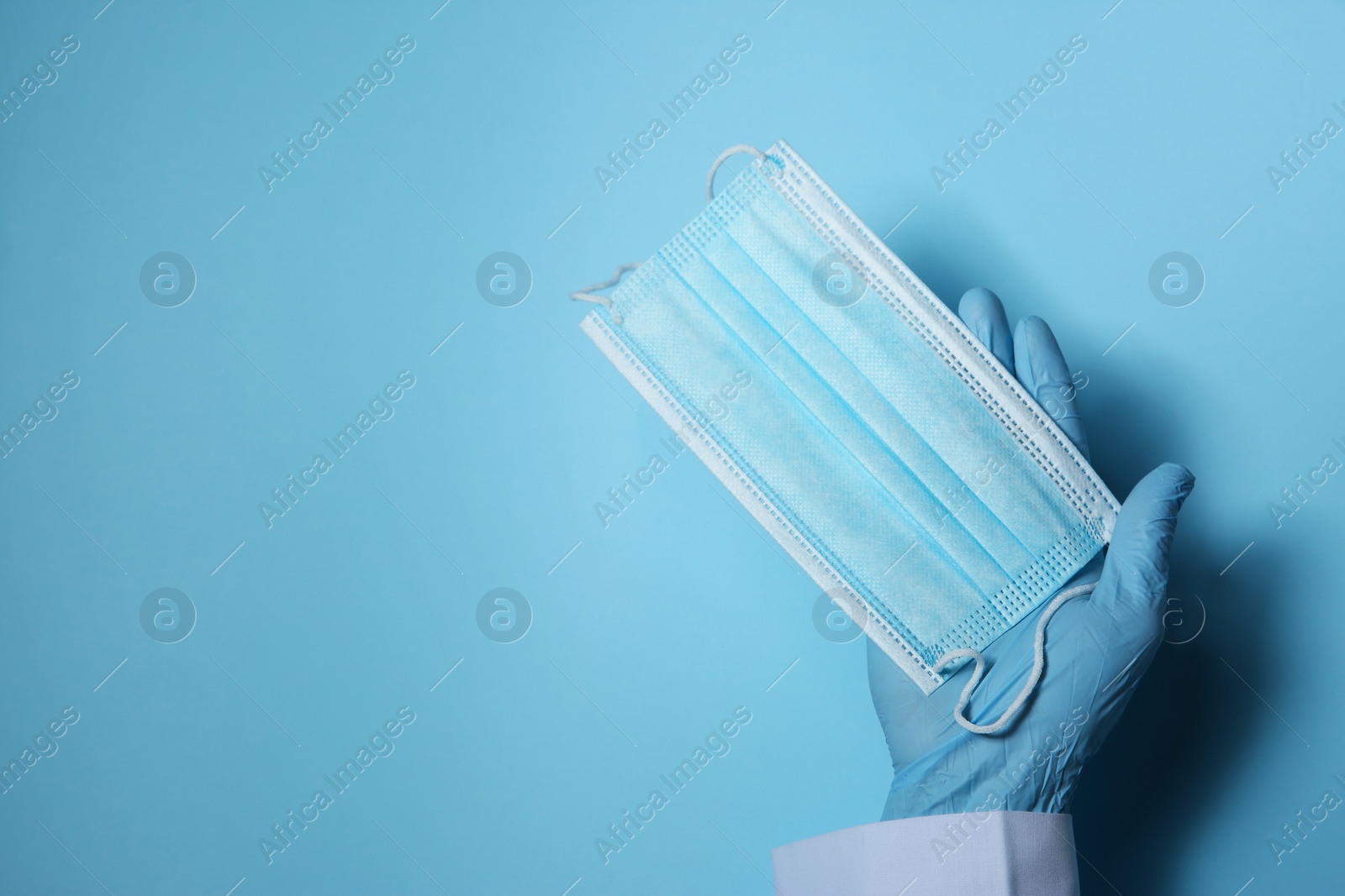 Photo of Doctor in latex gloves holding disposable face mask on light blue background, closeup with space for text. Protective measures during coronavirus quarantine